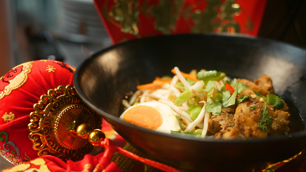 a bowl of meat, noodles and vegetables with an egg, surrounded by red Lunar New Year decorations.