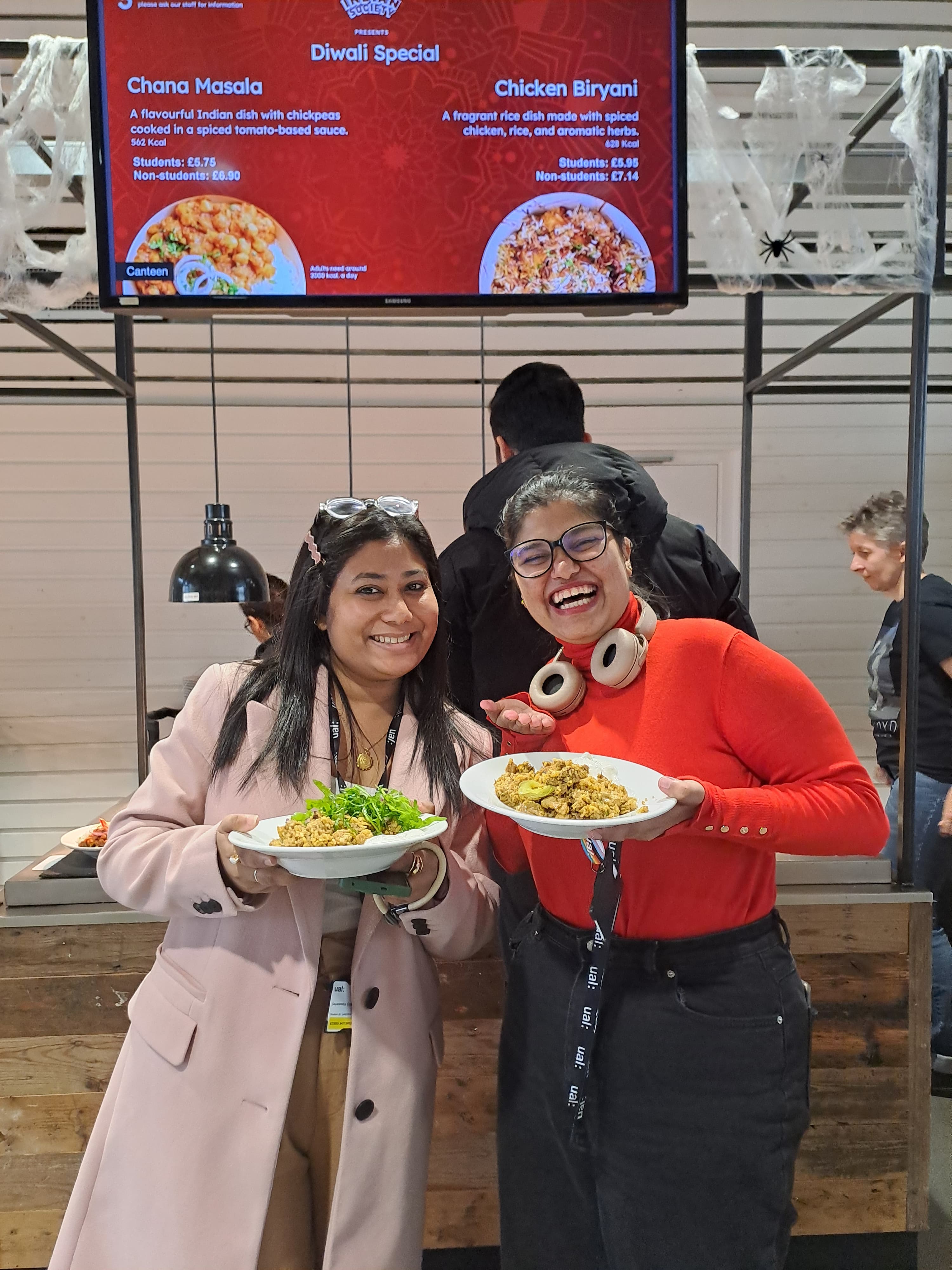 two people standing in front of a food counter smiling at the camera holding dishes full of food